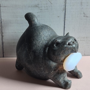 May include: A dark grey stone sculpture of a stout cat in a stretching pose with its tail pointed upward. The cat has small, dark, glossy eyes and is holding a smooth, translucent, pale white oval object in its mouth between its front paws.