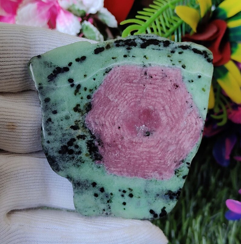 Puede incluir: Una piedra preciosa verde y rosa con inclusiones negras. La piedra preciosa tiene una forma hexagonal y est&aacute; rodeada de una matriz verde.