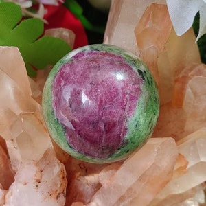 May include: A polished gemstone sphere, with a striking colour combination. The sphere has a central area of deep magenta, encircled by a green band with black speckles. The sphere rests on a bed of light orange crystals.