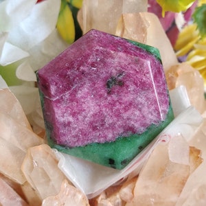 May include: A pink and green gemstone with a hexagonal shape. The gemstone is surrounded by white and beige crystals.