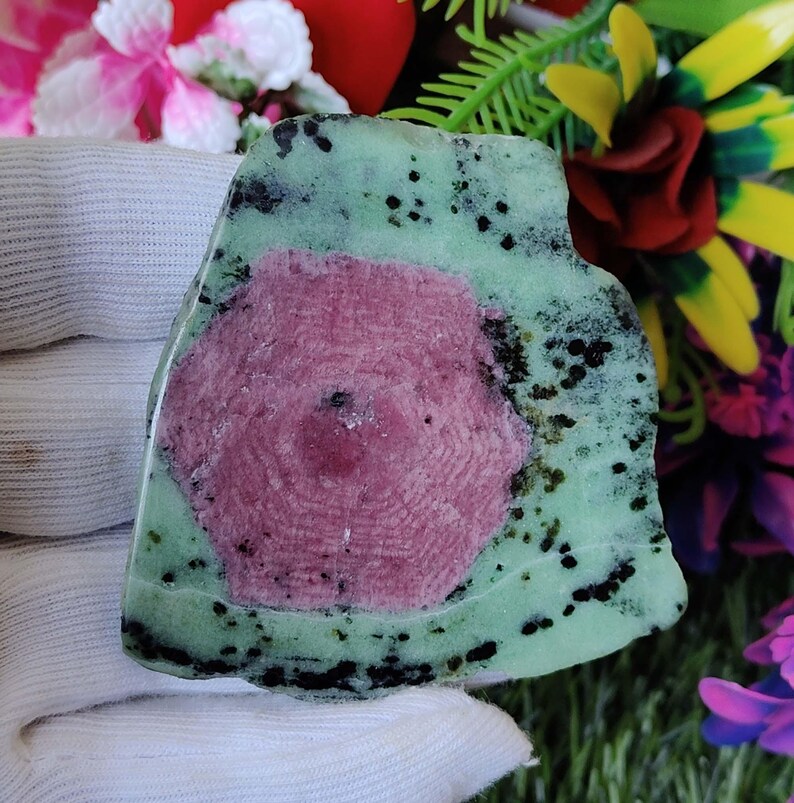Puede incluir: Una piedra preciosa verde y rosa con inclusiones negras. La piedra preciosa tiene un centro rosa circular con un patr&oacute;n radial y est&aacute; rodeada por un borde verde con manchas negras.