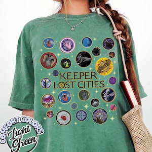 May include: Light green t-shirt with the text "Keeper of the Lost Cities" and various circular graphics. The shirt also features the words "Comfort Colors" and "Light Green" in a circular design.