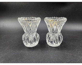 May include: Two clear, cut-glass candle holders. Each has a thistle-like shape with a round base, a textured body, and a jagged rim. The candle holders are displayed on a dark surface.