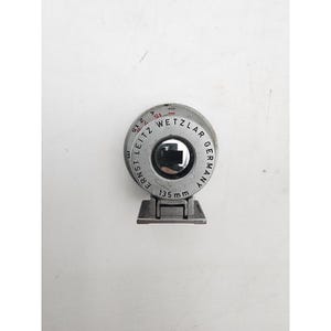 May include: A silver-toned metal camera viewfinder with a circular dial featuring the text ERNST LEITZ WETZLAR GERMANY and 135 mm. The device includes a rectangular mounting base and a central glass lens element with visible red and black markings.