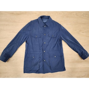 May include: A navy blue jacket with a collared neckline and long sleeves. The jacket has four front pockets and a button-up closure. The fabric has a slight sheen. The jacket is laid flat on a light wood surface.