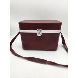 May include: A vintage brown rectangular case with a silver-colored trim and a secure latch. The case has a matching brown shoulder strap with a textured pad for comfort. The overall design suggests a classic travel or storage item.