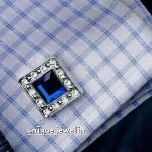 Puede incluir: Un gemelo de plata con una piedra preciosa azul cuadrada rodeada de piedras preciosas transparentes más pequeñas.