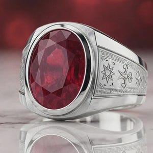 May include: A silver-toned ring featuring a large, oval-cut, deep red gemstone set in the center. The band is wide and polished, displaying intricate, engraved geometric patterns, including star and fleur-de-lis motifs along the sides.