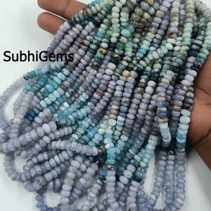 May include: Strands of small, round, multi-colored gemstone beads in shades of blue, gray, and green. The beads are strung together, creating a flowing, textured appearance. The text "SubhiGems" is visible in the lower left corner.
