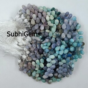 May include: A collection of multi-colored gemstone beads in shades of blue, green, and purple. The beads are teardrop-shaped and strung together. White tassels are visible on the left side of the image. The text "SubhiGems" is printed on the left side of the image.