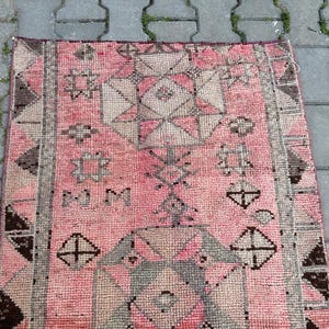 May include: A rectangular, hand-woven rug with a pink and gray geometric pattern. The rug features a central design of diamonds and stars, framed by a border of triangles and rectangles. The rug's color palette includes shades of pink, gray, and brown.