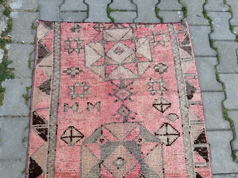 May include: A rectangular, hand-woven rug with a pink and gray geometric pattern. The rug features a central design of diamonds and stars, framed by a border of triangles and rectangles. The rug's color palette includes shades of pink, gray, and brown.