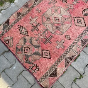 May include: A vintage, rectangular rug with a faded pink background and geometric patterns in beige, brown, and gray. The rug features a central medallion design and borders with repeating motifs, showcasing a traditional, distressed aesthetic.