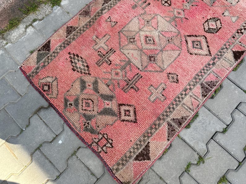 May include: A vintage, rectangular rug with a faded pink background and geometric patterns in beige, brown, and gray. The rug features a central medallion design and borders with repeating motifs, showcasing a traditional, distressed aesthetic.