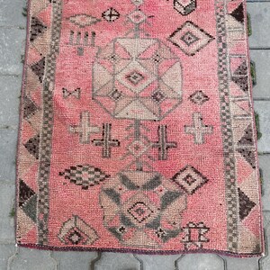 May include: A rectangular, hand-woven rug with a pink background and geometric patterns in shades of gray and beige. The rug features a central design with diamond and cross-like motifs, and a border with repeating shapes. This rug is suitable for home decor.