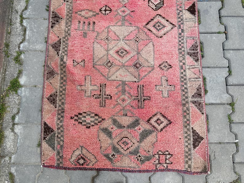 May include: A rectangular, hand-woven rug with a pink background and geometric patterns in shades of gray and beige. The rug features a central design with diamond and cross-like motifs, and a border with repeating shapes. This rug is suitable for home decor.