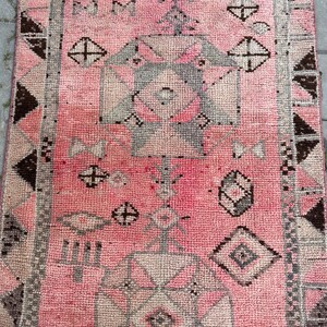 May include: A rectangular, pink rug with a geometric pattern. The rug features repeating shapes in shades of gray, brown, and pink. The design includes diamond, cross, and triangle shapes. The rug has a textured appearance.
