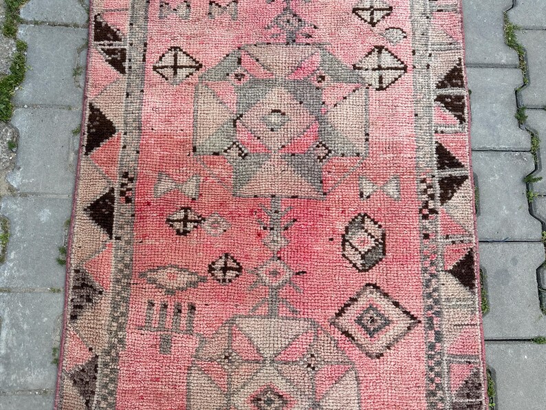 May include: A rectangular, pink rug with a geometric pattern. The rug features repeating shapes in shades of gray, brown, and pink. The design includes diamond, cross, and triangle shapes. The rug has a textured appearance.