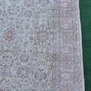 May include: A vintage rug with a light beige background and a brown floral pattern. The rug has a fringe border and is partially visible against a green background.