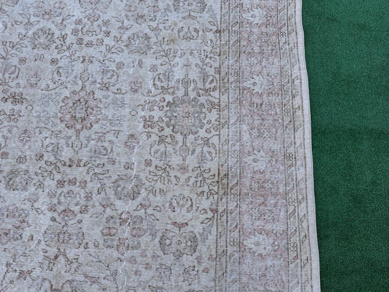May include: A vintage rug with a light beige background and a brown floral pattern. The rug has a fringe border and is partially visible against a green background.