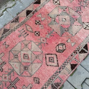 May include: A pink and gray patterned rug with geometric designs. The rug features a central field of pink with repeating geometric shapes in gray and beige. The border is a darker brown with geometric patterns. The rug is rectangular and appears to be made of woven material.