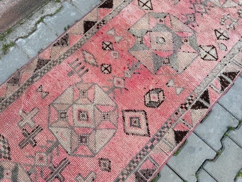 May include: A pink and gray patterned rug with geometric designs. The rug features a central field of pink with repeating geometric shapes in gray and beige. The border is a darker brown with geometric patterns. The rug is rectangular and appears to be made of woven material.