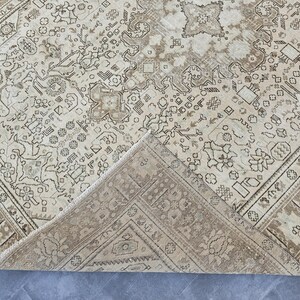 K&ouml;nnte beinhalten: Beige und brauner Vintage-Teppich mit aufwendigen geometrischen und floralen Mustern. Der Teppich hat einen Used-Look, mit einer gefalteten Ecke, die die Unterseite zeigt. Das Design zeigt ein zentrales Medaillon und Bord&uuml;rendetails.