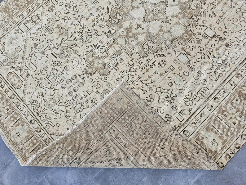 K&ouml;nnte beinhalten: Beige und brauner Vintage-Teppich mit aufwendigen geometrischen und floralen Mustern. Der Teppich hat einen Used-Look, mit einer gefalteten Ecke, die die Unterseite zeigt. Das Design zeigt ein zentrales Medaillon und Bord&uuml;rendetails.