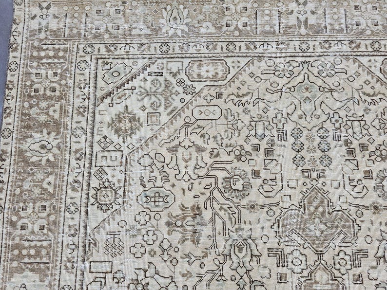 K&ouml;nnte beinhalten: Ausschnitt eines beigefarbenen und braunen Vintage-Teppichs mit einem aufwendigen, sich wiederholenden geometrischen Muster. Der Teppich zeigt ein zentrales Design mit floralen und abstrakten Formen, umrahmt von einem detaillierten Rand. Die Farbpalette ist neutral, mit Beige-, Braun- und Gr&uuml;nt&ouml;nen.