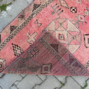 May include: A pink and brown vintage rug with geometric patterns. The rug has a distressed look, with faded colors and visible wear. The design includes diamond, cross, and square shapes. The rug is folded over, revealing the underside, which is a darker shade of brown.