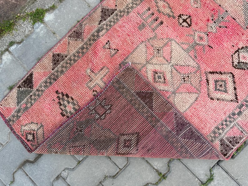 May include: A pink and brown vintage rug with geometric patterns. The rug has a distressed look, with faded colors and visible wear. The design includes diamond, cross, and square shapes. The rug is folded over, revealing the underside, which is a darker shade of brown.