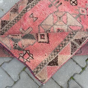May include: A folded, pink and brown rug with geometric patterns. The rug has a textured surface and a dark purple border. The design includes shapes like diamonds, triangles, and squares. The rug is placed on a gray, brick-like surface.