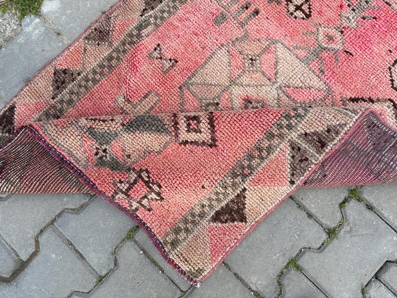 May include: A folded, pink and brown rug with geometric patterns. The rug has a textured surface and a dark purple border. The design includes shapes like diamonds, triangles, and squares. The rug is placed on a gray, brick-like surface.