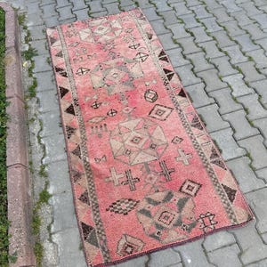 May include: A rectangular, vintage-style rug with a faded pink background and geometric patterns in beige, brown, and black. The rug features diamond, cross, and triangle motifs, and is placed on a brick surface.