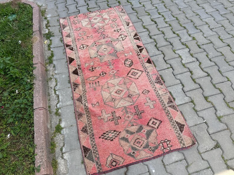 May include: A rectangular, vintage-style rug with a faded pink background and geometric patterns in beige, brown, and black. The rug features diamond, cross, and triangle motifs, and is placed on a brick surface.