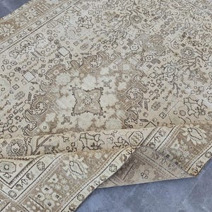 K&ouml;nnte beinhalten: Beige-brauner Vintage-Teppich mit einem komplizierten Blumen- und geometrischen Muster. Der Teppich hat einen Used-Look, mit einer aufgerollten Kante, die die Unterseite zeigt. Die Farbpalette umfasst Beige-, Braun- und Cremet&ouml;ne und erzeugt eine warme &Auml;sthetik.