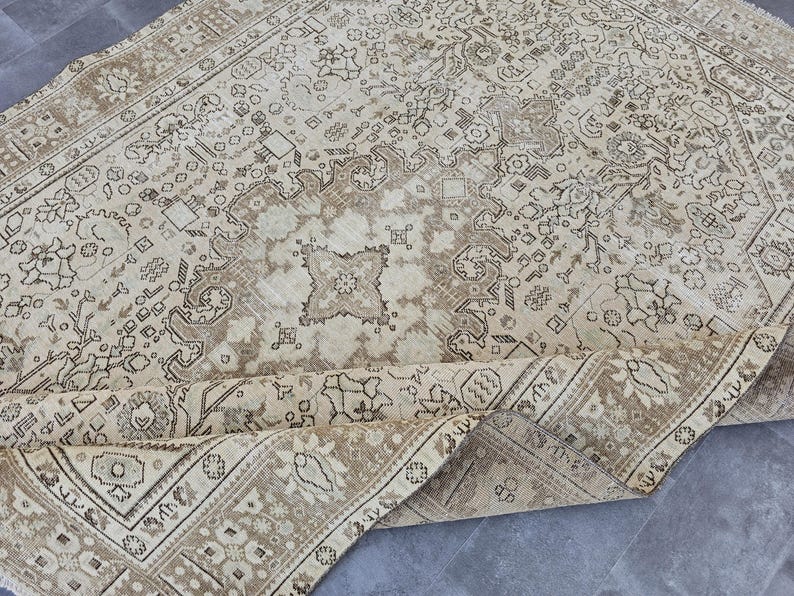 K&ouml;nnte beinhalten: Beige-brauner Vintage-Teppich mit einem komplizierten Blumen- und geometrischen Muster. Der Teppich hat einen Used-Look, mit einer aufgerollten Kante, die die Unterseite zeigt. Die Farbpalette umfasst Beige-, Braun- und Cremet&ouml;ne und erzeugt eine warme &Auml;sthetik.
