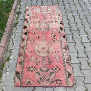 May include: A rectangular, vintage rug with a faded pink background and geometric patterns in shades of beige, brown, and black. The rug features diamond, cross, and other abstract shapes. The rug is placed on a gray brick surface.