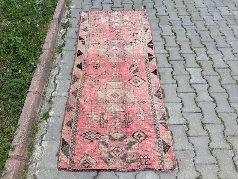 May include: A rectangular, vintage rug with a faded pink background and geometric patterns in shades of beige, brown, and black. The rug features diamond, cross, and other abstract shapes. The rug is placed on a gray brick surface.