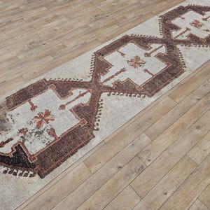 Puede incluir: Una alfombra de pasillo blanca con un patrón geométrico marrón y rojo. La alfombra tiene un aspecto desgastado y es perfecta para añadir un toque de estilo bohemio a tu hogar.