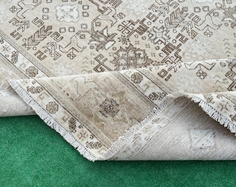 Può includere: Tappeto vintage beige e marrone con intricati motivi geometrici. Il tappeto presenta un bordo dettagliato e un design centrale con motivi floreali e astratti. Il tappeto è parzialmente arrotolato, rivelando il bordo sfrangiato e il lato inferiore.