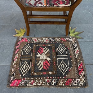 May include: A small, brown and black patterned rug with a pink and white border. The rug has a geometric design with a central motif.