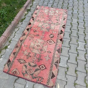 May include: A rectangular, vintage-style rug with a faded red and brown color palette. The rug features geometric patterns, including diamonds, crosses, and other shapes. The rug is placed on a gray brick surface next to a concrete border with green grass.