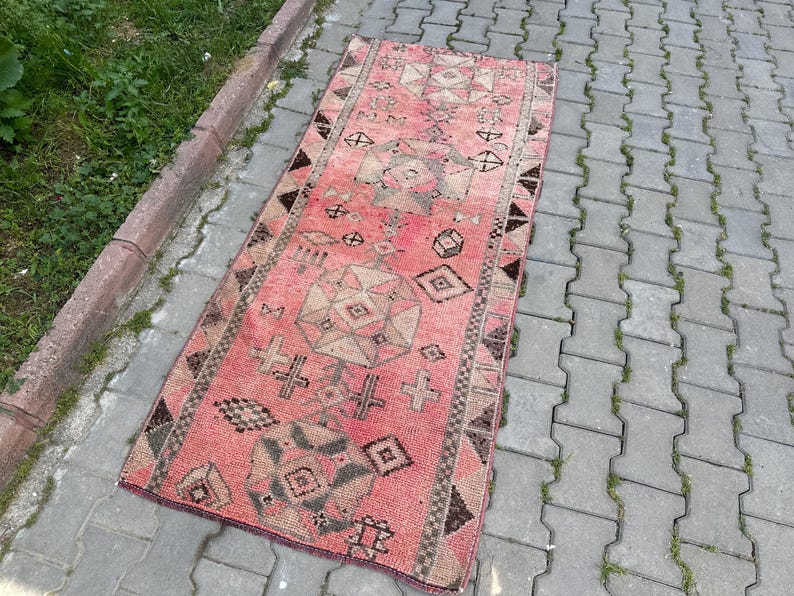 May include: A rectangular, vintage-style rug with a faded red and brown color palette. The rug features geometric patterns, including diamonds, crosses, and other shapes. The rug is placed on a gray brick surface next to a concrete border with green grass.