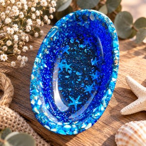 Könnte beinhalten: Eine ovale Schale aus Kunstharz in tiefem Königsblau mit eingebetteten blauen Seesternen, glitzernden Partikeln und zerkleinerten Muschelfragmenten am Rand. Die Innenfläche ist glatt und glänzend und zeigt ein dichtes, funkelndes, vom Ozean inspiriertes Design.