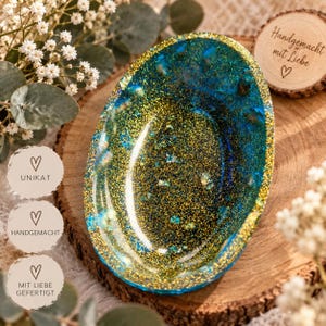 Könnte beinhalten: Eine ovale Schmuckschale aus Kunstharz mit einer lebendigen Mischung aus tiefblauem Farbton und schimmerndem Goldglitter. Die Schale hat eine glatte, glänzende Oberfläche mit einem markanten goldfarbenen Rand, der das Licht reflektiert.