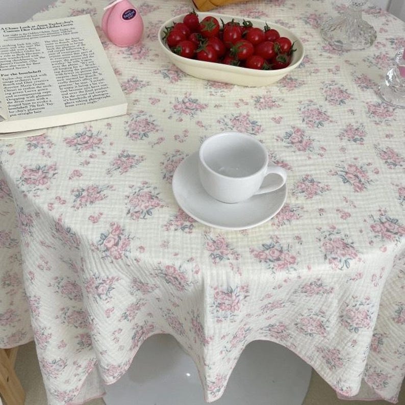 May include: A white tablecloth with a pink and green floral pattern, featuring a white ceramic teacup and saucer, a bowl filled with red cherry tomatoes, a pink spherical object, and an open book with printed text on the surface.