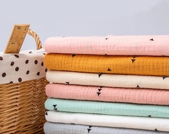 May include: A stack of colorful muslin fabric in shades of pink, yellow, cream, and blue-green, each with a small black triangle pattern. The fabric is stacked next to a wicker basket with a polka-dot lining.