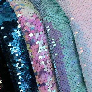 May include: A collection of fabric swatches featuring dense, overlapping circular sequins in a gradient of colours. The textures transition from deep metallic blue to iridescent pink, light purple, and a pale, shimmering white finish.