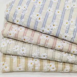 May include: Four folded pieces of fabric featuring a repeating pattern of white daisy flowers with yellow centers. Each piece has a different colored vertical stripe background in light blue, dusty pink, sage green, and mustard yellow.